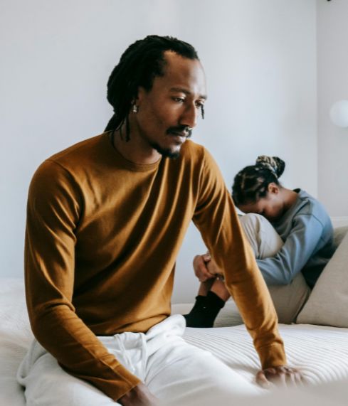 A man sitting on the edge of a bed, looking away, while his partner sits curled up behind him.